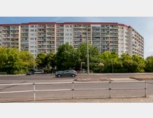 Eine große Wohnanlage mit mehreren Etagen, Balkonen und farbigen Fassadenelementen, umgeben von Bäumen und Grünflächen. Im Vordergrund verläuft eine Straße mit parkenden und fahrenden Autos sowie einem Metallgeländer entlang der Gehwegkante.