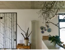 Wohnraum mit minimalistischer Dekoration: Eine weiße Wand mit Baumtapete auf der linken Seite und blauen Ziertellern auf der rechten Seite. Ein Zweig mit Lichtkugeln hängt von der Decke herab, daneben ein Tisch mit einer Vase und Kerzenständern sowie eine Zimmerpflanze auf einer weißen Ablage.