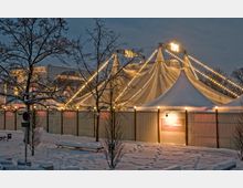 Ein großes Zirkuszelt mit angestrahlten Lichtgirlanden steht in einer verschneiten Winterlandschaft, umgeben von einem Holzzaun. Im Vordergrund sind schneebedeckte Bäume, Sitzbänke und Gehwege zu sehen.