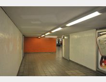 Unterführung mit gefliesten Wänden und rechteckigen Deckenleuchten, bestehend aus einem orangefarbenen Abschnitt und weißen gefliesten Wänden; im Hintergrund sind eine Tür sowie Rolltreppen sichtbar.