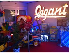 Ein dekorierter Raum mit einem roten Teppich, einem beleuchteten Schriftzug "Gigant", einem Glücksrad, Spielautomaten und einer Mini-Kinderfahrzeug-Attraktion. Im Hintergrund befinden sich bayerische Elemente wie blau-weiße Muster und ein Dekoelement mit einem lachenden Gesicht.