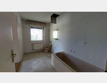 Ein leeres Badezimmer mit weißen Wandfliesen und beigem Bodenfliesen. Es enthält eine braune Badewanne, eine Toilette, ein Fenster mit Rolladen sowie einen Heizkörper unterhalb des Fensters.