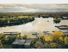 Luftaufnahme eines Flusses mit angrenzendem Wald und herbstlich gefärbten Bäumen. Im Vordergrund befinden sich ein Gebäude, kleinere Bootsanleger mit mehreren Booten und Frachtschiffe, während im Hintergrund eine Stadtlandschaft zu sehen ist.
