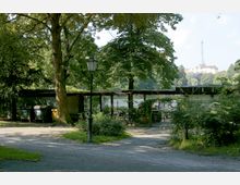 Außenbereich eines Parks mit einem kleinen Pavillon, umgeben von Bäumen und Sträuchern. Im Hintergrund sind ein See und ein Funkturm erkennbar, sowie ein Gebäude auf einer Anhöhe.