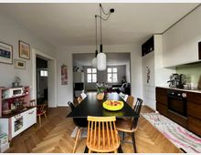 Wohnküche mit modernem Design: ein schwarzer Esstisch mit verschiedenen Holzstühlen steht im Vordergrund, darüber hängt eine minimalistische Deckenleuchte. Im Hintergrund sind eine offene Wohnzimmerecke mit Sofa und ein heller Holzboden zu sehen, außerdem rechts eine Küchenzeile mit weißen Oberschränken, dunklen Unterschränken und einer Kaffeemaschine.