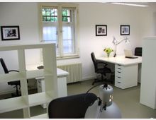 Modernes Büro mit weißen Möbeln, darunter Arbeitstische, Rollcontainer und ein Regal. Im Raum befinden sich schwarze Bürostühle, eine Zimmerpflanze, Schreibtischlampen sowie ein Fenster mit geteilten Scheiben und Blick auf Grünflächen.