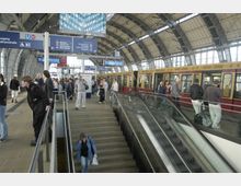 Ein Bahnhof mit modernem, gewölbtem Glasdach, mehreren Gleisen und einer einfahrenden S-Bahn in den Farben Rot und Gelb. Menschen stehen auf dem Bahnsteig, während einige die Treppen und Rolltreppen nutzen.