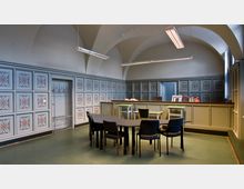 Ein Raum mit hohen, gewölbten Decken und dekorativen Wandpaneelen mit floralen Mustern. In der Mitte stehen ein schwarzer Tisch und mehrere Stühle; im Hintergrund befindet sich ein Tresen mit Büromaterialien.