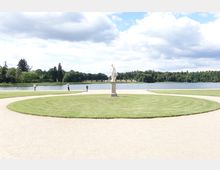 Eine Parklandschaft mit einem runden, gepflegten Rasenbereich im Vordergrund, in dessen Zentrum eine Statue steht. Im Hintergrund ist ein See mit umliegenden Bäumen und einem leicht bewölkten Himmel zu sehen.