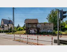 Ehemaliges Bahnhofsgebäude in Werder (Havel) mit beschädigtem Dach und beschmierten Wänden, umgeben von Gleisen, einer Straße und grüner Vegetation. Im Hintergrund stehen Wohnhäuser und Bäume, während ein Bahnübergang im Vordergrund sichtbar ist.
