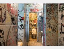 Öffentliche Toilette mit Wänden und Türen, die vollständig mit Graffiti, Schriftzügen und Zeichnungen bedeckt sind. Der Boden besteht aus hellen Fliesen, und ein WC mit offenem Deckel steht im Fokus des Bildes.