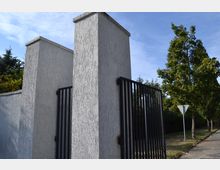 Zementierte Eingangssäulen mit grauer Verputzoberfläche und einem schwarzen Metallzaun. Im Hintergrund sind grüne Bäume, ein Gehweg und ein Verkehrsschild zu sehen.
