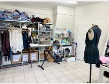 Raum einer Schneiderei mit Regalen voller Stoffe, Kleidungsstücken und Nähzubehör; eine Schneiderpuppe mit einem schwarzen Kleid steht im Vordergrund. Im Hintergrund befinden sich eine Waschmaschine, Reinigungsmittel und ein Bügelbrett.