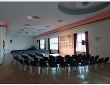 Ein Veranstaltungssaal mit Holzboden, einer kleinen Bühne mit Mikrofonen und Stühlen, die halbkreisförmig angeordnet sind. Große Fenster mit roten Vorhängen und Kunstwerke an der Wand prägen den Raum.