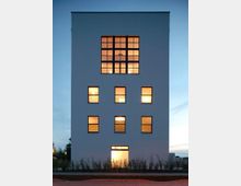 Moderne Gebäudefassade mit weißem Putz und rechteckigen Fenstern, die im oberen Bereich eine große Fensterfront bilden. Das Gebäude ist von niedrigen Pflanzen umgeben und wird bei Dämmerung von innen beleuchtet.