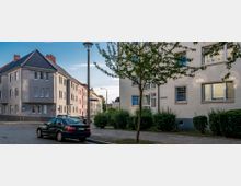 Straßenszene in einem Wohngebiet mit mehrstöckigen, pastellfarbenen Wohnhäusern. Einzelne geparkte Autos und ein Gehweg mit Bäumen und Hecken prägen das Bild, während eine Straßenlaterne in der Mitte der Szene steht.