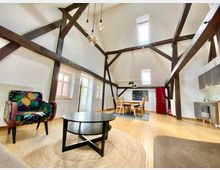 Großzügiger Wohnraum in einem offenen Dachgeschoss mit sichtbaren dunklen Holzbalken, weißen Wänden und Holzboden. Der Raum ist eingerichtet mit einem bunten Sessel, einem runden Couchtisch, einem Esstisch mit Stühlen, einer kleinen Küchenzeile und einem roten Kühlschrank.