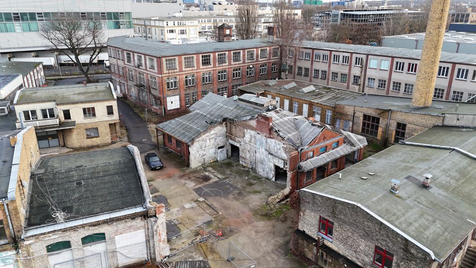 Luftaufnahme eines alten Fabrik- und Industrieareals mit mehreren Backstein- und Putzgebäuden, teils beschädigten Dächern und einem offenen Innenhof mit Betonflächen. Rechts ragt ein hoher Schornstein neben langen Werkhallen hervor, im Hintergrund stehen weitere größere Gewerbebauten.
