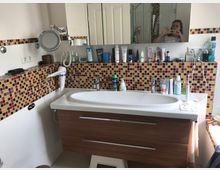 Modernes Badezimmer mit Waschtisch in Holzoptik, rechteckigem Waschbecken und großem Wandspiegel. Die Wand ist mit weißen Fliesen und einem Mosaikmuster in Rot-, Gelb- und Brauntönen gestaltet, mit zahlreichen Hygiene- und Pflegeprodukten auf der Ablage.