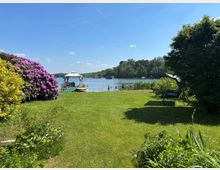Grüne Rasenfläche am Ufer eines Sees mit Steg, kleinem Boot und weißer Überdachung. Links blühender Strauch, rechts Bäume und eine Gartenschaukel; gegenüber ein bewaldetes Ufer unter blauem Himmel.