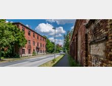 Straßenszene mit historischen roten Backsteingebäuden auf beiden Seiten, teils verfallen, mit Graffiti an den Wänden. Die Szenerie enthält einen Bürgersteig, eine Straße, Bäume und einen klaren Himmel mit weißen Wolken.