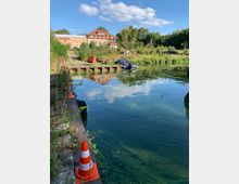 Teich mit grünem Wasser und Vegetation am Ufer, im Hintergrund ein ziegelrotes Gebäude mit Dachfenstern und grüne Bäume. Im Vordergrund befinden sich Baukegel, Holzstege und ein mit einer Plane abgedecktes kleines Boot.