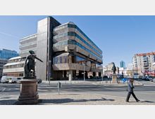 Modernes Verwaltungsgebäude mit kantiger Architektur und großen Fenstern, flankiert von zwei historischen Statuen auf Sockeln. Im Vordergrund eine gepflasterte Straße und Gehwege, im Hintergrund urbane Bebauung mit modernen und älteren Gebäuden.