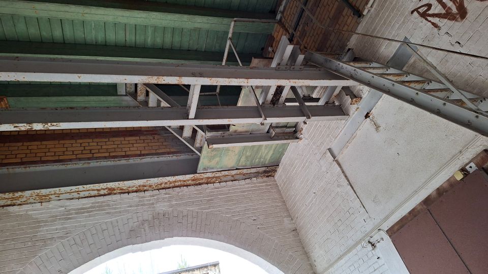 Blick nach oben in einen industriellen Innenraum mit weiß gestrichenen Ziegelwänden und einem Rundbogen, darüber grün gestrichene Holzdecke. Rostige Stahlträger, Metallstreben und eine Leiterkonstruktion verlaufen entlang der Wand, daneben ist ein großes Metalltor zu sehen.