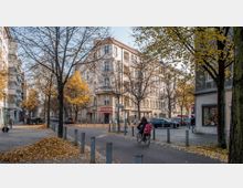 Straßenszene in einer urbanen Umgebung mit Herbstlaub auf dem Boden und kahlen Bäumen. Ein Eckgebäude im Gründerzeitstil mit Geschäften im Erdgeschoss dominiert das Bild, während Personen zu Fuß und ein Fahrradfahrer die Szene beleben.
