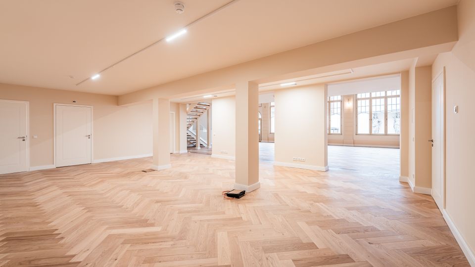 Großer, leerer Innenraum mit hellem Fischgrätparkett, beigen Wänden und mehreren Stützsäulen sowie Decken-Spotbeleuchtung. Im Hintergrund sind eine offene Treppe und große Sprossenfenster zu sehen, die viel Tageslicht in den Raum lassen.
