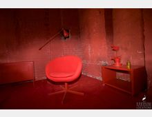 Roter Raum mit roten Ziegelwänden, einem roten Teppichboden, einem modernen roten Sessel, einem roten Beistelltisch mit Topfpflanze und Dekorationsobjekt sowie einer roten Stehlampe.