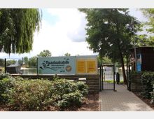 Eingang zu einer Flussbadestelle mit einem Weg, einem Metalltor und einem Schild mit der Beschriftung „Flussbadestelle Nationalparkstadt Schwedt/Oder e. V.“. Im Hintergrund sind Bäume, ein Fluss und Teile eines Stegs sichtbar, umgeben von üppigem Grün.