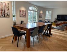 Wohnzimmer mit hellem Fischgrätenparkett, einem großen Holztisch mit unterschiedlichen Stühlen, einer Sitzecke mit Sofa und großem Fernseher sowie dekorativen Wandbildern. Große Fenster und eine Doppeltür mit Rundbogen lassen Tageslicht in den Raum.