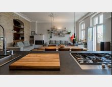 Offene Wohnung mit moderner Küche und Wohnzimmer in einem stilvollen Design; im Vordergrund eine Küchentheke mit Schneidebrett und Gasherd, dahinter ein Essbereich mit Holztisch und Stühlen, ein großes Sofa, eine Ziegelwand mit integriertem Regal und ein Wandkamin. Große Fenster und eine Balkontür auf der rechten Seite sorgen für viel Tageslicht.