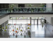 Großer, moderner Innenraum mit minimalistischem Design, bestehend aus einer unteren Ebene mit Tischen und bunten Stühlen sowie einer Galerieebene mit schwarzen Stühlen und Pflanzen. Große Glasfronten geben Blick auf den Außenbereich frei.