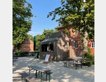 Holzhütte in einem Biergarten mit Kiesboden, umgeben von Bäumen und roten Backsteingebäuden. Mehrere Holzbänke und Tische stehen im Außenbereich, ergänzt durch Aufsteller mit Tafeln.