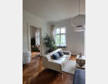 Wohnzimmer mit hellen Wänden, einem hölzernen Fischgrätparkettboden, einem cremefarbenen Ledersofa, einem Couchtisch aus Naturstein, einer großen Zimmerpflanze und Fenstern mit Rundbogen. Im Hintergrund ist ein angrenzender Raum mit einem Schreibtisch sichtbar.