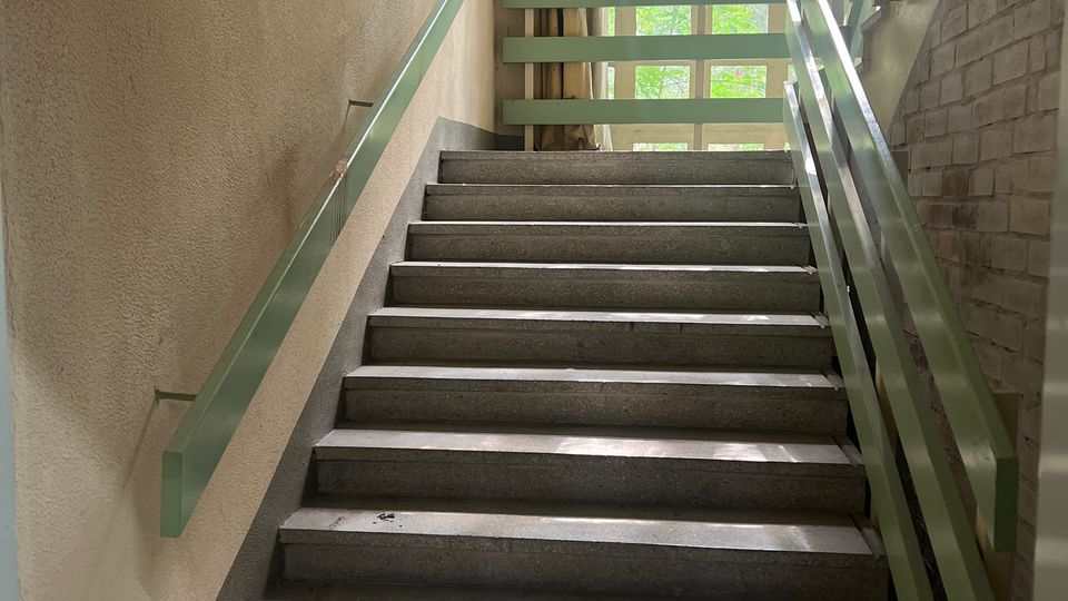 Innen-Treppenhaus mit breiten grauen Steinstufen und beidseitigen hellgrünen Metallgeländern. Am oberen Ende sorgt ein großes Rasterfenster für Tageslicht; rechts eine Ziegelwand, links eine verputzte Wand mit Vorhang.