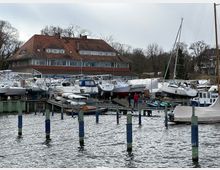 Ein kleiner Hafen mit mehreren an Land abgestellten Motor- und Segelbooten, teilweise mit Abdeckungen. Im Hintergrund ein großes Gebäude mit rotem Ziegeldach, umgeben von Bäumen und winterlicher Landschaft.