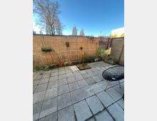 Kleiner, gepflasterter Hinterhof mit Sichtschutzwand aus Schilfrohrmatten und vereinzelten Pflanzenkübeln. Im Vordergrund steht ein modern geformter Stuhl, im Hintergrund sind kahle Bäume und ein klarer blauer Himmel zu sehen.