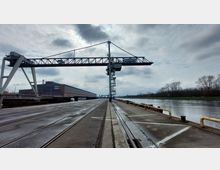 Industriegelände mit einem Hafenkai entlang eines Flusses. Im Vordergrund sind Schienen und eine große, über dem Kai montierte Kranganlage zu sehen, im Hintergrund ein Lagergebäude mit der Aufschrift „MEERA REMINE“.