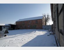 Verschneiter Innenhof eines Bauernhofs mit einer großen roten Backsteinscheune mit grünen Holztoren und wenigen kleinen Fenstern. Rechts begrenzt ein verwittertes Wirtschaftsgebäude den Hof, im Hintergrund stehen ein Metalltor und ein kahler Baum vor klarem Himmel.