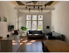 Wohnzimmer mit hellen Wänden und Holzfußboden, ausgestattet mit zwei grauen Sofas, einem runden Holztisch und einem niedrigen weißen Sideboard mit Fernseher. Große Fenster lassen Tageslicht herein, eine Zimmerpflanze steht auf einem kleinen Tisch, und eine Hängelampe mit mehreren Glühbirnen ist an der Decke montiert.