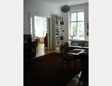 Helles Wohnzimmer mit Holzboden, großem Teppich, einem Esstisch mit Stühlen und einer grauen Couch vor Fenstern, die auf eine grüne Außenansicht blicken. Im Hintergrund befindet sich ein Bücherregal und eine offene Verbindung zu einem weiteren Raum mit Sitzmöbeln und großen Fenstern.