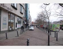 Straßenszene in einer Stadt mit Kopfsteinpflaster, flankiert von Gebäuden mit Geschäften wie einem Brillenatelier. Auf der rechten Seite stehen Bäume ohne Laub und ein grüner Lebensmittelanhänger, im Hintergrund sind Marktstände und ein Pkw sichtbar.