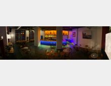 Eine moderne Bar mit Sitzbereich, in warmem und farbigem Licht beleuchtet. Der Raum enthält runde Tische mit Stühlen, eine große Theke mit Regalen voller Getränkeflaschen im Hintergrund und dekorative Elemente wie Pflanzen und Wandbilder.