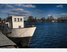 Ein weißes Boot liegt an einem Metallsteg vor Anker in einem ruhigen Gewässer. Im Hintergrund sind Uferbäume, ein Schornstein und Liegeplätze mit Booten zu sehen, unter einem Himmel mit vereinzelten Wolken.