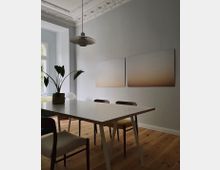 Moderner Essbereich in einem hellen Raum mit Holzboden und Stuckdecke. Ein weißer Tisch mit unterschiedlichen Stühlen steht vor einer Wand mit zwei abstrakten Gemälden, daneben eine große Fensterfront mit Blick ins Grüne.