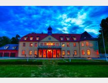 Zweistöckiges Herrenhaus mit gelbem Putz und rotem Satteldach, beleuchtet durch orangefarbenes Licht aus den Fenstern, vor einem abendlichen Himmel mit dichten blauen Wolken. Im Vordergrund befindet sich eine gepflegte Rasenfläche.