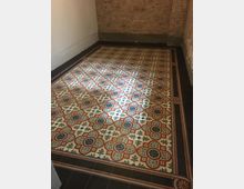 Ein kleiner Raum mit unverputzten Ziegelwänden und einem bunt gemusterten Boden aus dekorativen Fliesen mit floralen Motiven in Rot, Blau und Beige. Der Fliesenbereich ist von einem dunklen Rand mit geometrischen Elementen eingefasst.